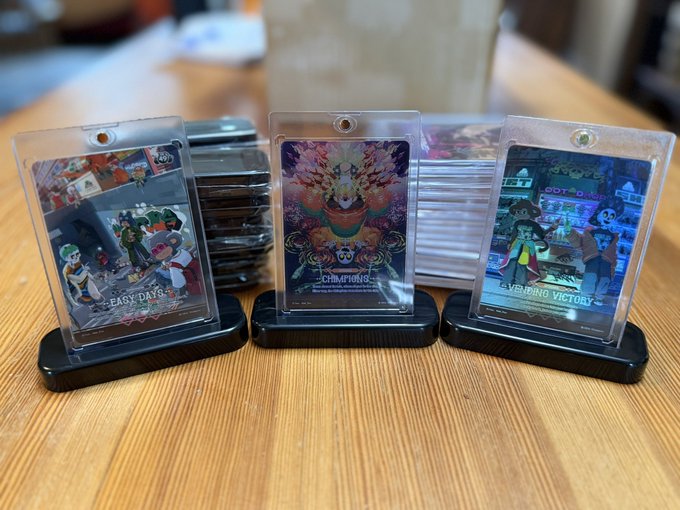 Three display stands on a wooden table holding what appear to be game cards or collectibles in clear cases, with labels including 'EASY DAYS', 'CHAMPIONS', and 'GRINDING VICTORY' visible.