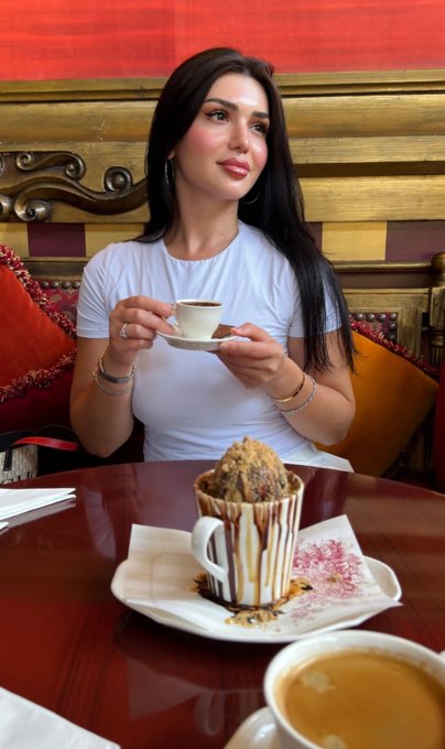 A woman with long dark hair enjoys a relaxed Sunday morning, sipping from a small espresso cup while a decadent dessert sits on the polished wooden table in front of her. The warm red tones and ornate woodwork in the background complement the cozy "Gm gm Sunday" vibe of leisure and indulgence.