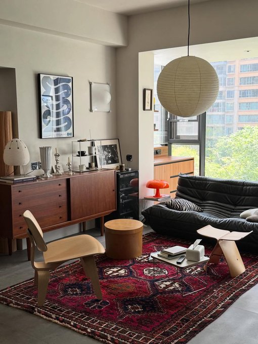 Stylish mid-century modern living room interior featuring a black Togo sofa, wooden Eames-style chair, paper lantern, and Persian rug.