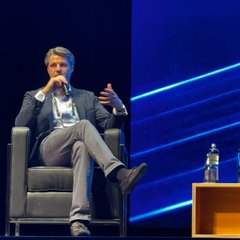 A man sits cross-legged on a stage holding a microphone and gesturing with one hand, with a water bottle nearby and blue lighting behind him.