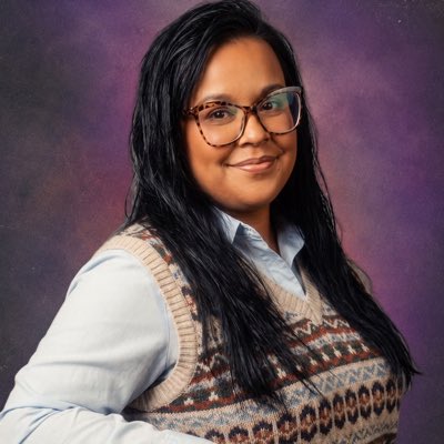 A studio portrait of a smiling woman with long dark hair and glasses, wearing a light blue shirt under a patterned sweater vest against a purple backdrop.