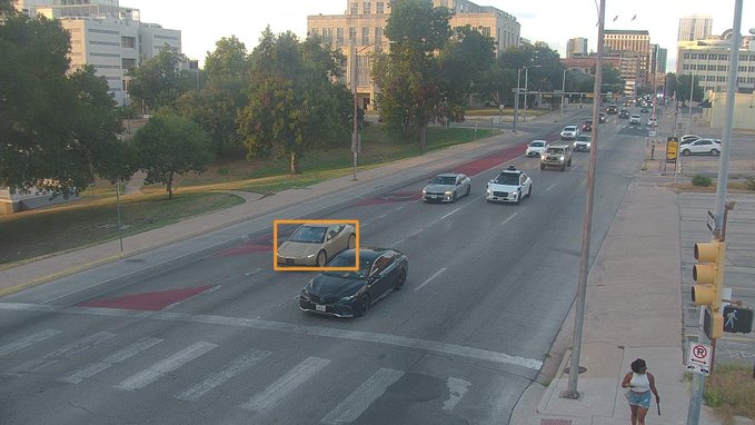 A street scene at an intersection showing several cars driving on a multi-lane road, with a beige car highlighted by an orange bounding box.