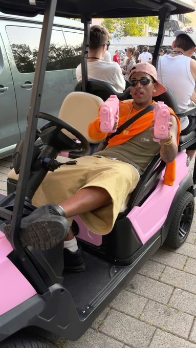 A man in an orange shirt, khaki shorts, sunglasses, and a peach cap sits in a pink golf cart holding two pink toy guns, with a crowd and white vehicles visible in the background.
