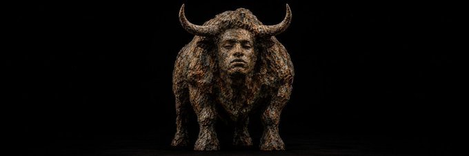 A dark, textured sculpture of a bull with a human-like face stands against a black background.