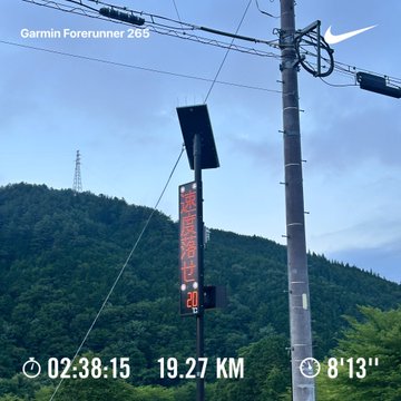 A Garmin Forerunner 265 smartwatch screen displays a run of 19.27 km completed in 2:38:15 with an average pace of 8'13", set against a backdrop of a forested mountain landscape and a digital road sign. The image visually captures the post's morning run stats and reflects the achievement of completing nearly the full 19 km on foot, with the mountain scenery echoing the beautiful summit view mentioned.