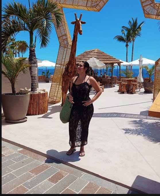 A woman in a black lace dress stands at a tropical resort near a large giraffe statue, with palm trees, beach umbrellas, and the ocean visible in the background.