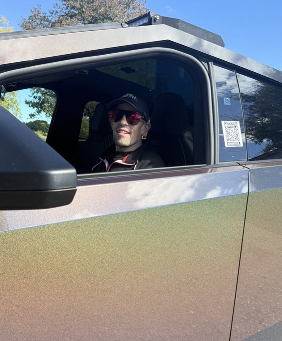 A person wearing sunglasses and a cap smiles from the driver's seat of a dark vehicle parked outdoors on a sunny day, with a QR code displayed on the window.