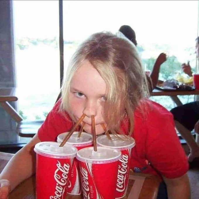 A blonde woman using four straws simultaneously to drink from four large Coca-Cola cups clumped together on a table.