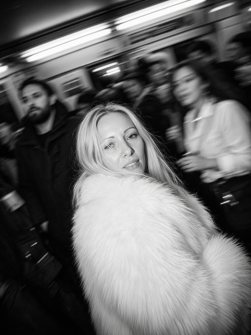 A black and white street photography portrait of a blonde woman in a fur coat looking at the camera, with motion-blurred people in the background.