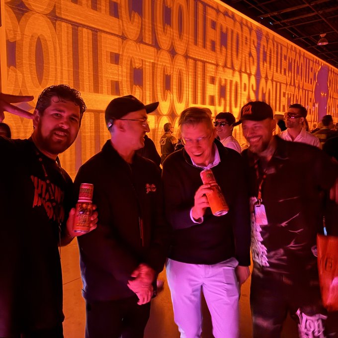 Four men pose with bottles in a red-lit event space, with a large background sign repeatedly displaying “COLLECTORS COLLECTORS.”