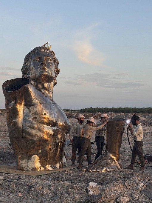 A group of workers in a rocky desert landscape assemble a large golden metallic sculpture of a woman with a crown, while one worker uses a blowtorch on a foot piece.