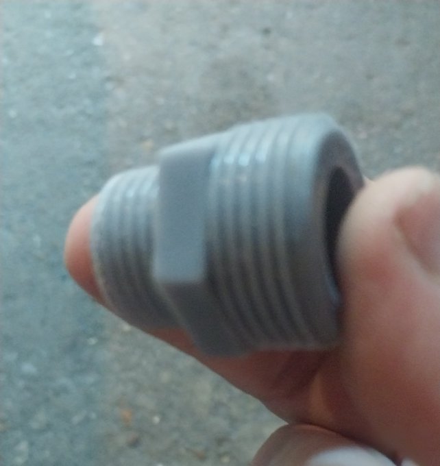 A close-up, slightly blurry photo of a hand holding a gray threaded PVC pipe fitting (hex bushing) against an asphalt ground background.