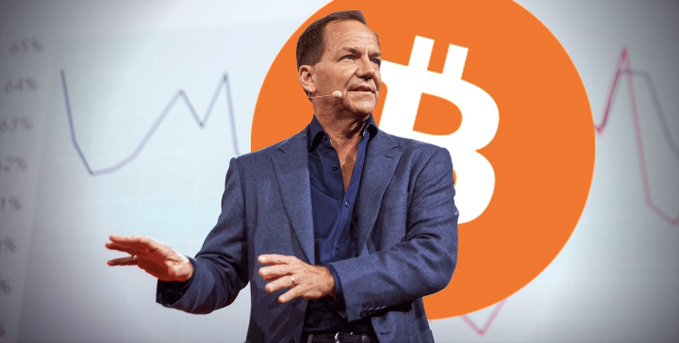Paul Tudor Jones speaks on stage in front of a large Bitcoin logo, reflecting the news that his Tudor Investment firm has increased its holdings in BlackRock's IBIT Bitcoin ETF to 688,529 shares worth $22.9 million.