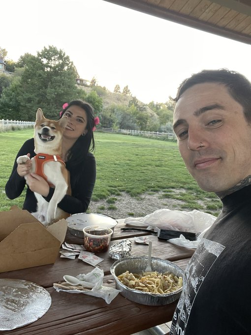 A couple poses for a selfie at a picnic table outdoors; the woman holds a smiling Shiba Inu puppy in a red harness while the man films them. Food containers are scattered on the table.
