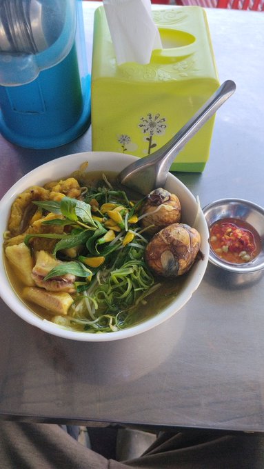 The image shows a bowl of food sitting on top of a table with a green box next to it. The bowl contains noodles, meat, and vegetables such as broccoli and carrots. A spoon is also present near the bowl. In addition to this, there are two bowls in the scene - one containing soup and another possibly holding more food items or condiments.