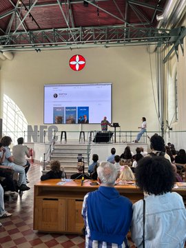 The image shows a group of people gathered around a large screen displaying information about the @NFC Summit held at the Lisbon Convention Center. The attendees are seated, with some standing near the stage where a speaker is giving a presentation. A few individuals can be seen holding handbags and backpacks as they listen to the event. The atmosphere appears to be one of learning and engagement, with people actively participating in discussions or taking notes on their devices.