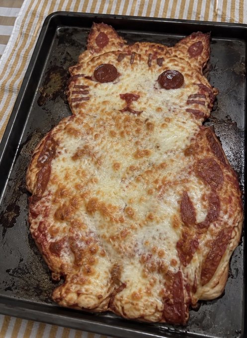 A homemade pizza shaped like a cat with pepperoni eyes and whiskers on a baking tray.
