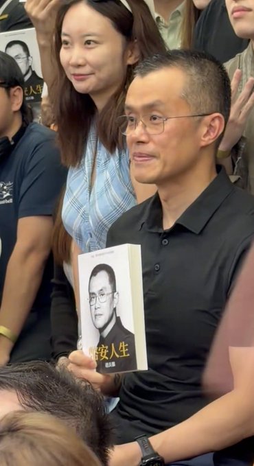 A man with glasses wearing a black polo shirt holds up a book with his portrait on the cover, with Chinese text '人生' (Life) visible, surrounded by people in the background.