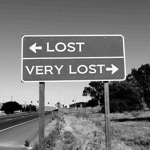 A black and white photo of a road sign reading '← LOST' on top and 'VERY LOST →' on the bottom, standing beside a road.