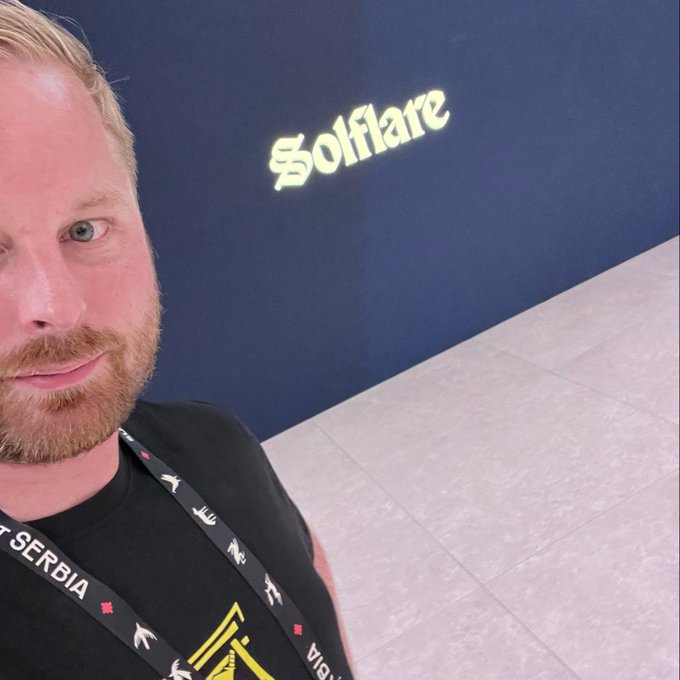 A selfie of a bearded man wearing a black shirt and lanyard, taken in front of a dark wall displaying a glowing 'Solflare' logo.