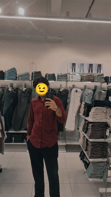 A person wearing a red button-up shirt and black pants taking a mirror photo in what looks like a clothing store, face obscured by an emoji.