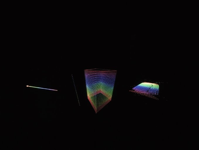 The image displays a dark void featuring three colorful, abstract geometric forms: a thin diagonal line on the left, a vertical shield-like shape with a rainbow gradient in the center, and a multicolored triangular plane on the right. These appear to be visualizations of Kim's pixel-based digital artworks, illustrating the original digital assets that collectors were paying to have others fly to Palo Alto to mint on their behalf due to the location-restricted minting process.