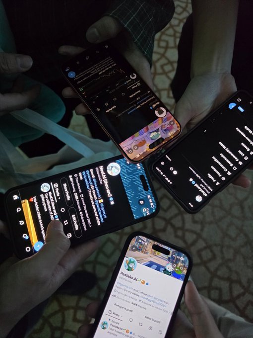 Four people huddle together in a circle, each holding up their smartphone screens displaying crypto-related social media posts, with one device showing "Pasteke.lol" and various other feeds related to trading content, capturing a moment of shared discussion among the group.