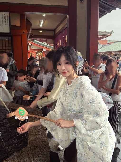 A young woman in a traditional floral kimono and decorative hairpiece uses a wooden ladle at a Japanese temple purification fountain, participating in the ritual of washing hands before prayer—a gesture of seeking good fortune. The "BC.GAME" logo is overlaid on the image, connecting the temple blessing to the sponsor's wish for luck.