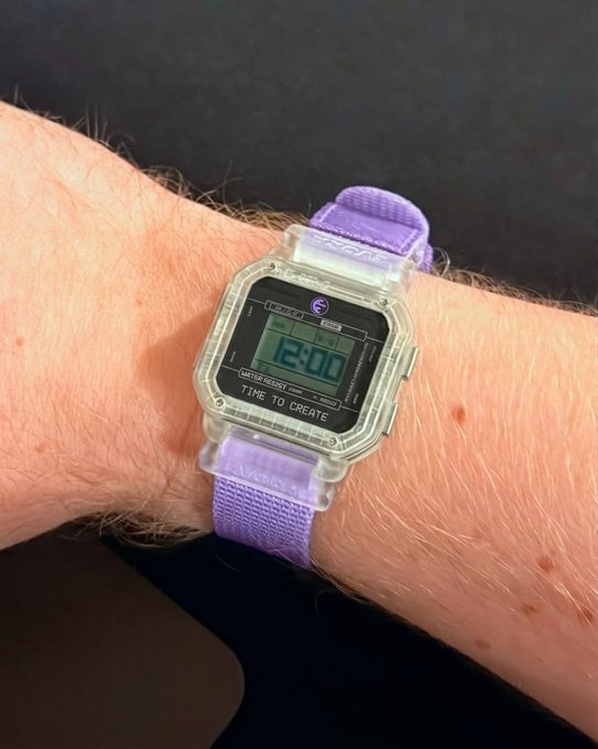 A close-up of a wrist wearing a transparent digital watch with a purple velcro strap displaying '12:00' and the text 'TIME TO CREATE'.