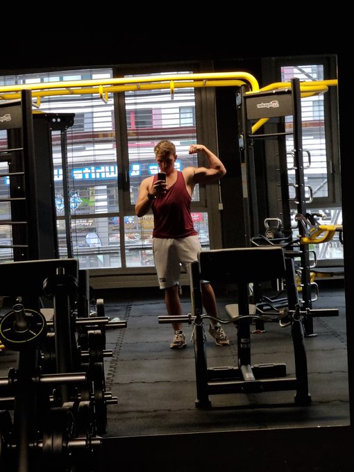 A man in a red tank top and grey shorts flexes his bicep while taking a mirror selfie with his phone in a gym.