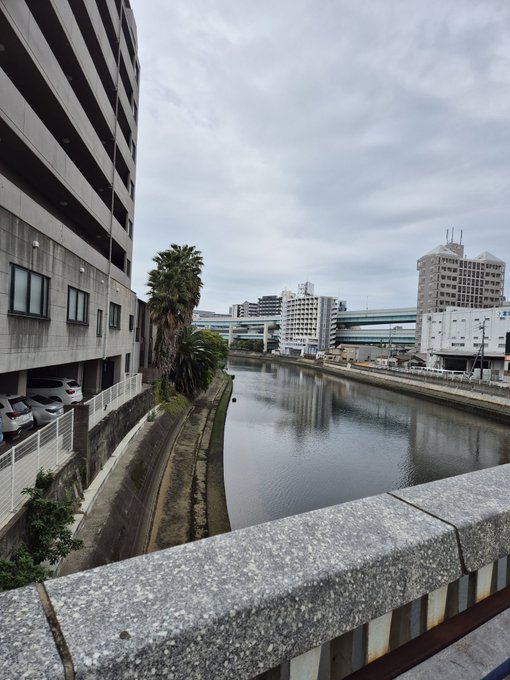 A view of a city river or canal with apartment buildings and a train track in the background, taken from a concrete bridge.