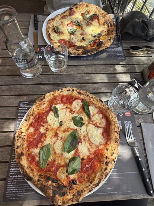 Two wood-fired pizzas served on white plates on a wooden table with placemats—one appears to be a margherita with fresh basil and the other topped with vegetables and cheese—accompanied by glasses of water and cutlery.