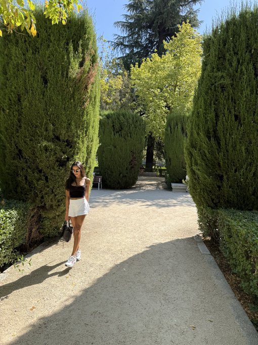 A young woman in a black top, white skirt, and sunglasses walks along a gravel path lined with tall, manicured cypress trees and other greenery.