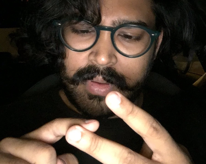A man with glasses, a beard, and curly dark hair wearing a black shirt, with his hands raised near his face.