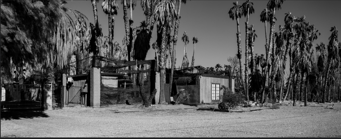 A black and white panoramic photograph shows an abandoned building surrounded by tall palm trees.
