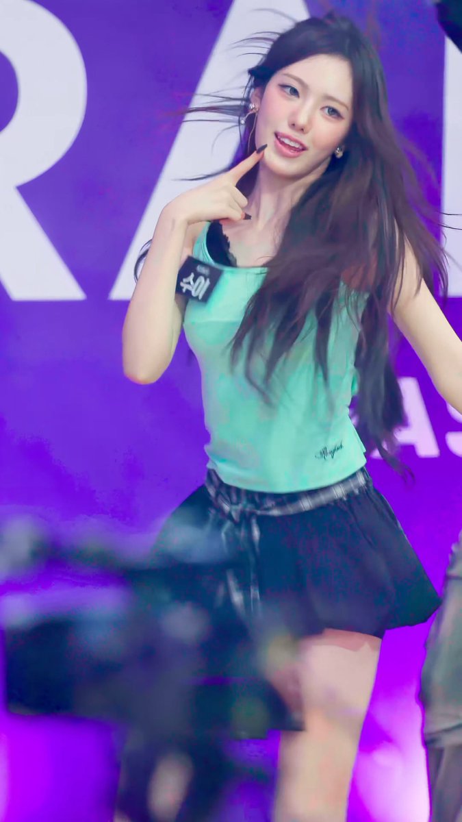 A performer on stage poses with her finger to her cheek, wearing a mint green tank top and black pleated skirt, with purple stage lighting in the background.