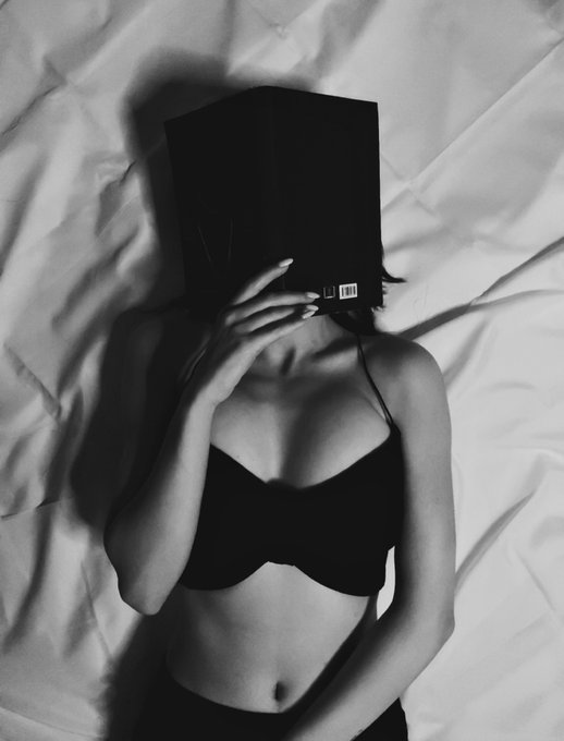 Black and white photo of a woman in a black bra lying on white sheets, holding a book over her face, with only her torso and hand visible.