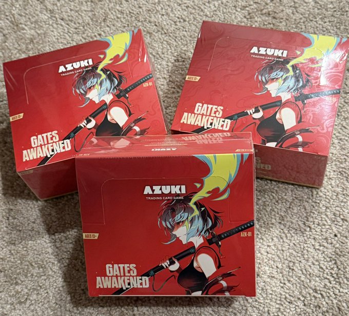 Three sealed boxes of the "AZUKI Trading Card Game" - "Gates Awakened" ATK-01 set, each featuring anime character artwork, displayed on a carpeted floor—marking the start of a new Azuki trading card collection.