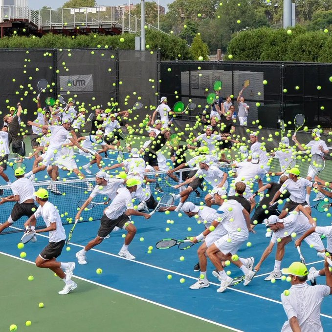 A large group of tennis players in white outfits and neon caps are actively hitting tennis balls on a blue court during what appears to be a chaotic group tennis event, with balls flying everywhere.