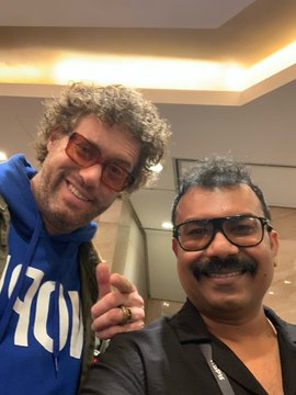 Selfie of two smiling men; one with curly hair wearing orange-tinted sunglasses and a blue sweatshirt, the other with dark hair, a mustache, and black glasses wearing a dark jacket.