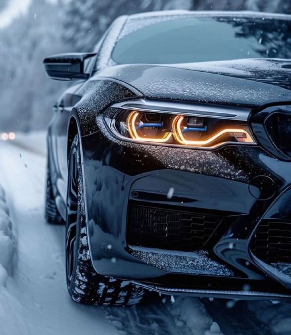 A close-up photograph of a black BMW with distinctive orange angel-eye headlights parked on a snowy road.