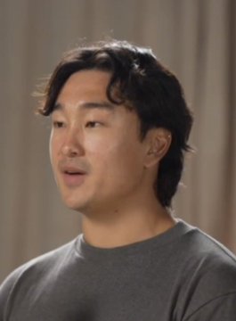 An East Asian man with wavy dark hair, wearing a gray shirt, is captured mid-speech in an interview setting.