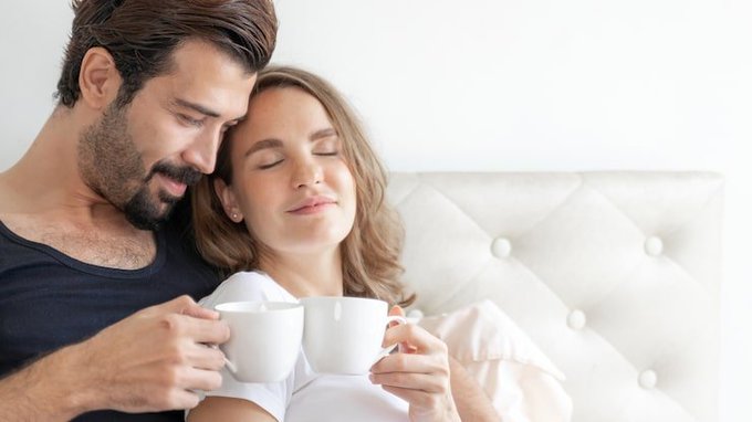 A couple shares an intimate moment while drinking from white ceramic mugs, with a bearded man resting his head against a smiling woman with closed eyes on a tufted white couch.