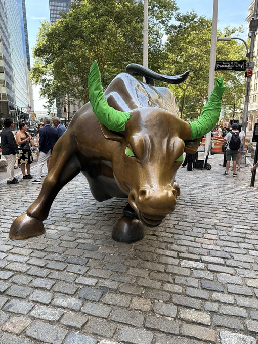 The Charging Bull statue, with its horns wrapped in green, stands prominently on a cobblestone plaza near Bowling Green, symbolizing bullish market strength. The visible "Evacuation Bay" sign and crowds in the background frame this iconic symbol of financial optimism—mirroring the post's message about the unstoppable "Golden Bull Movement" behind $Gayu.