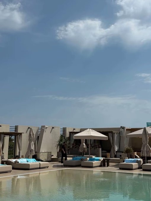 A poolside scene at a resort featuring a swimming pool in the foreground, white sun loungers with blue towels, beige umbrellas, white cabanas, and a person standing near the pool under a bright blue sky with scattered clouds.