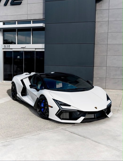 A white Lamborghini Revuelto is parked outside a modern building marked '5135' on a sunny day.