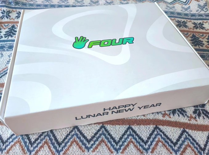 The image features a white box with a green logo that says "Four" on it, placed on top of a brown blanket or rug. The box contains a gift for the new year, which appears to be related to sports. The text inside the box reads "Happy Lunar New Year", and there is also an additional text that reads "Guess what's inside: A Sport". This suggests that the gift box was likely sent as a birthday present or a congratulatory gift for someone who enjoys sports.