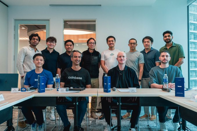 A group of people, with one wearing a 'coinbase' t-shirt, pose together behind a table in what appears to be a corporate or office setting.