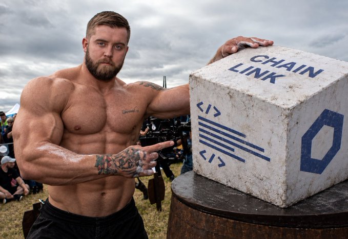 A muscular, shirtless bearded man with tattoos on his arms flexes and points at a large concrete block labeled 'CHAIN LINK' with a chain link logo, outdoors at what appears to be a fitness event.