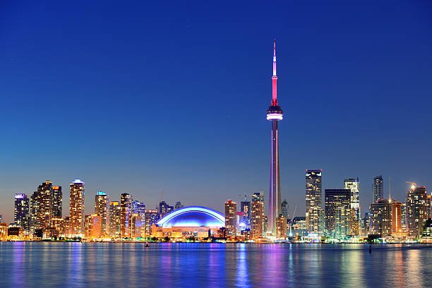 A nighttime skyline view of Toronto, Canada, featuring the illuminated CN Tower, the glowing dome of the Rogers Centre, and city lights reflecting on the water of Lake Ontario.